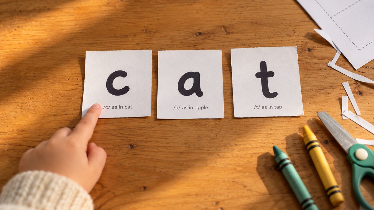 A child's finger pointing at printed letter cards c, a, t on a kitchen table